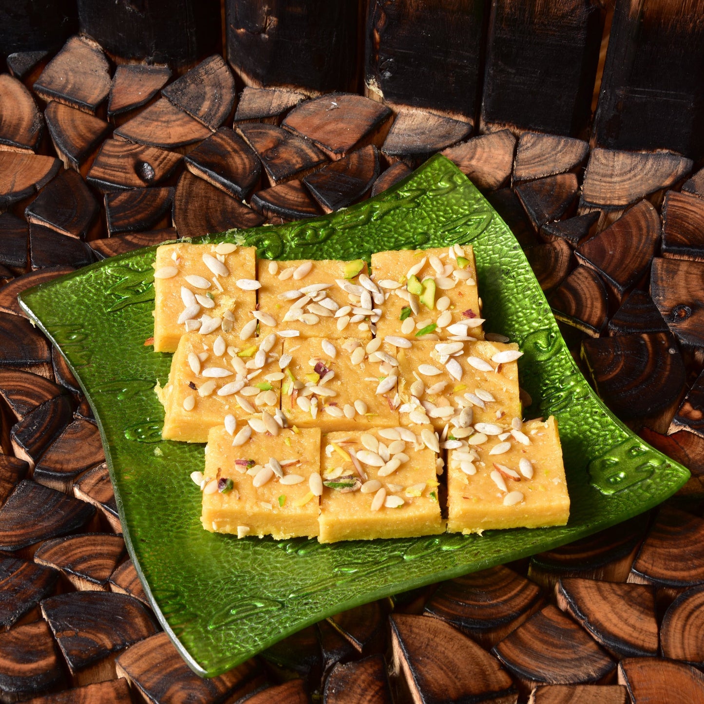 Square yellow barfis with white melon seeds on a green plate, placed on a wooden surface.